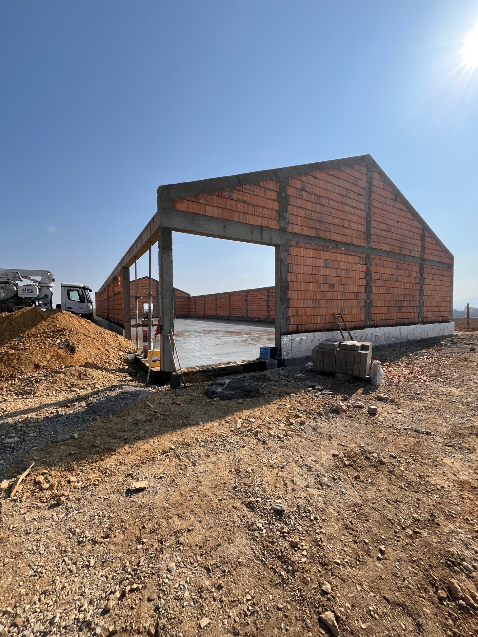 Budowa hali: ściany z czerwonej cegły, betonowy szkielet, wylana posadzka. Na zewnątrz hałda ziemi i betoniarka. Słoneczny dzień, błękitne niebo.