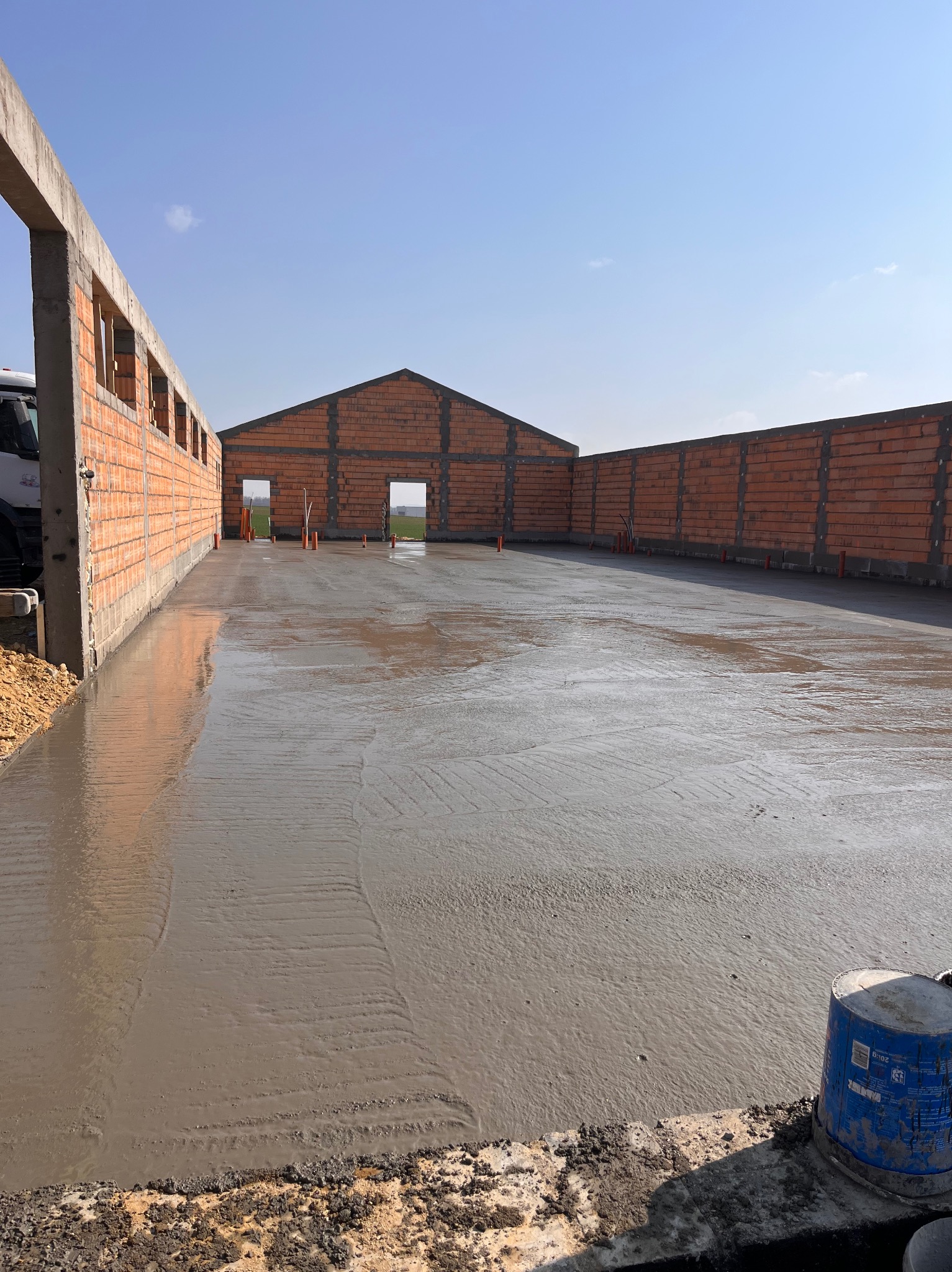 Świeżo wylana, gładka posadzka betonowa wewnątrz budynku w trakcie budowy, z widocznymi ścianami z czerwonej cegły i elementami instalacji. Jasne, naturalne światło.