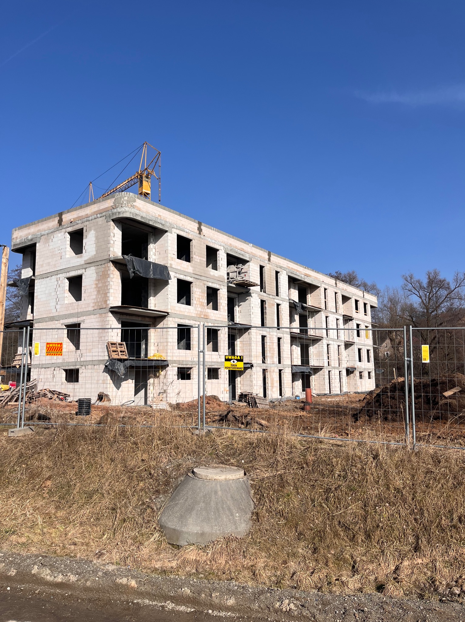 Trzykondygnacyjny budynek w stanie surowym otwartym, ogrodzony siatką, z widocznym żurawiem na dachu i błękitnym niebem w tle. Widoczne pustaki i otwory okienne.