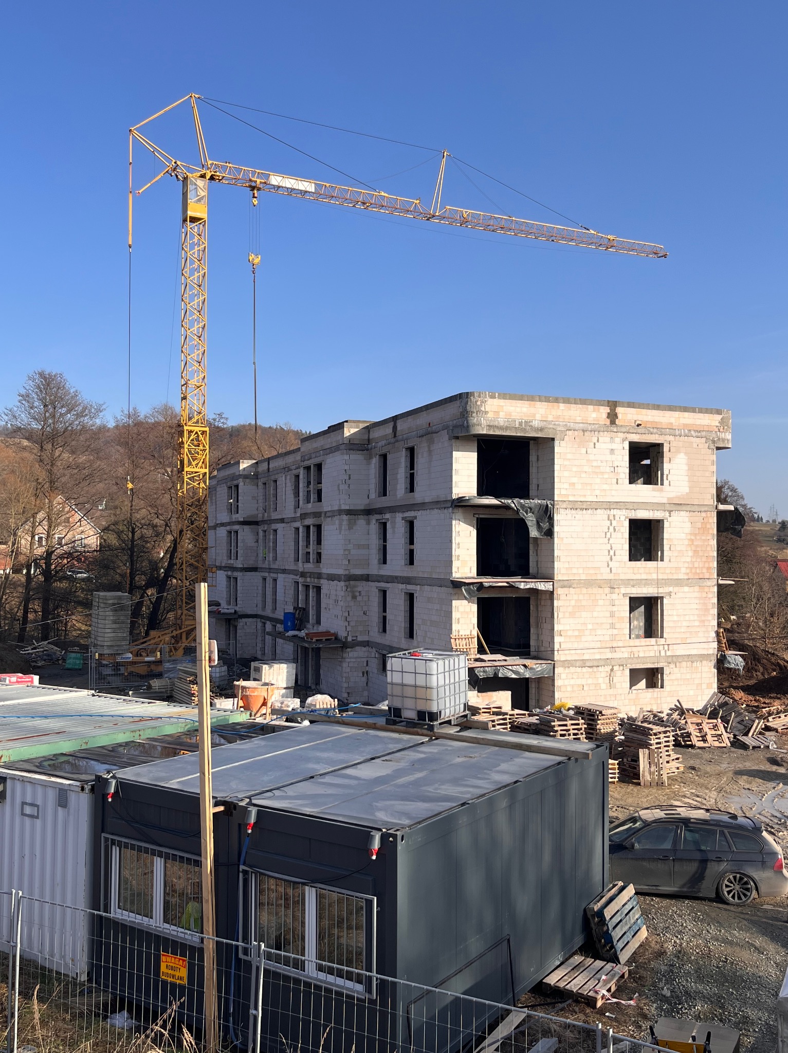 Budowa trzypiętrowego budynku z betonu komórkowego w trakcie realizacji, widoczny żuraw budowlany i kontenery biurowe. Słoneczny dzień, błękitne niebo.
