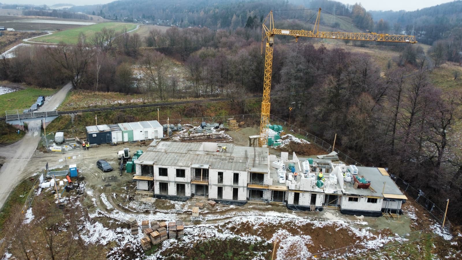Budowa dwukondygnacyjnego budynku z białego bloczka, widok z góry. Na dachu widoczne zbrojenia, w tle żuraw budowlany i zalesione wzgórza. Częściowo ośnieżony teren.
