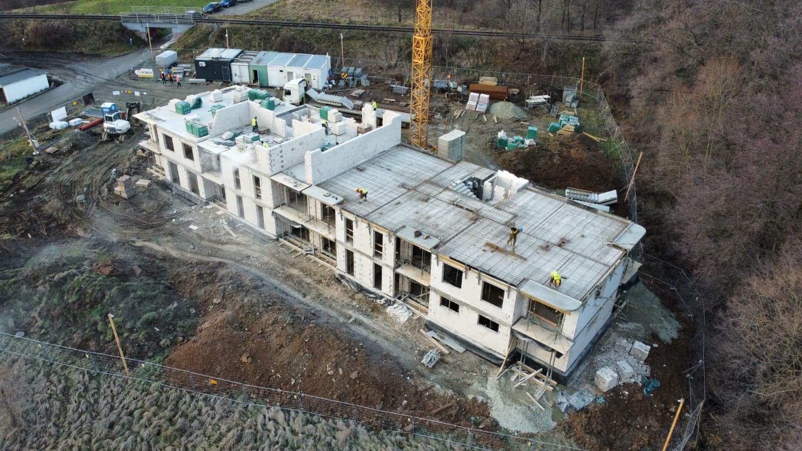 Wysoka perspektywa na dwukondygnacyjny budynek w trakcie budowy. Widoczne zbrojenia na dachu, ściany z pustaków i pracownicy w kamizelkach odblaskowych. Żuraw budowlany w tle.