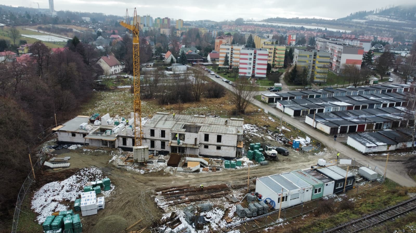 Budowa dwukondygnacyjnego budynku z pustaków, widok z góry. Na pierwszym planie składowane materiały budowlane, w tle zabudowa miejska i dźwig budowlany.