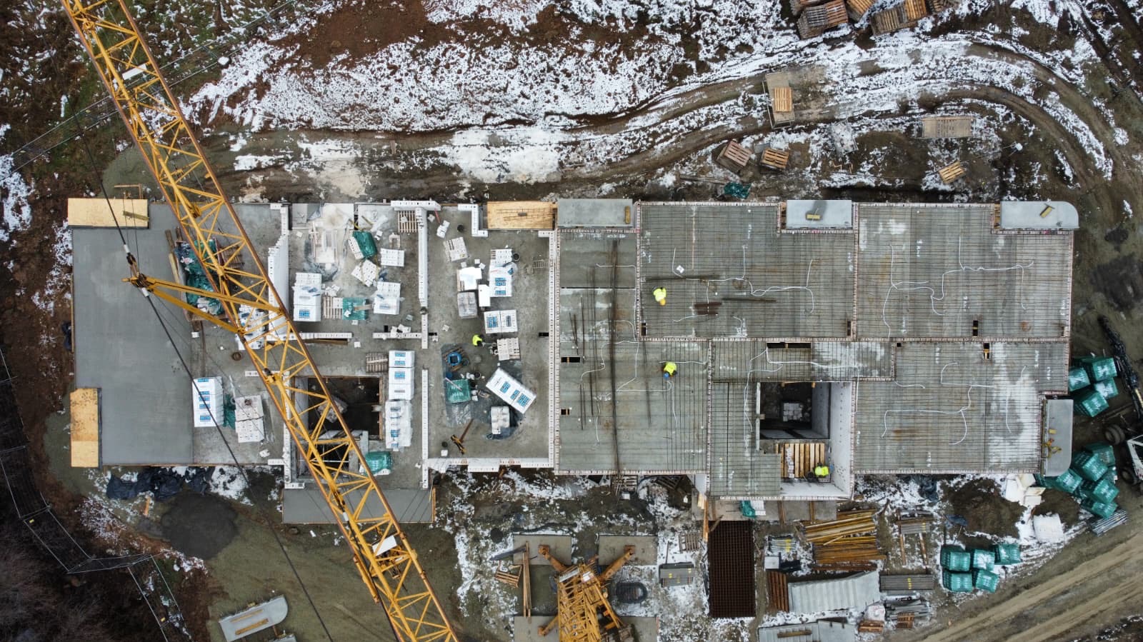 Widok z drona na plac budowy z fundamentami, częściowo zalanymi betonem, zbrojeniami i żółtym dźwigiem. W tle teren pokryty śniegiem.