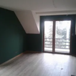 Pomalowane na ciemnozielono wnętrze poddasza z drewnianą podłogą imitującą panele, widoczne okno balkonowe i dwuskrzydłowe okno z widokiem na zewnątrz.