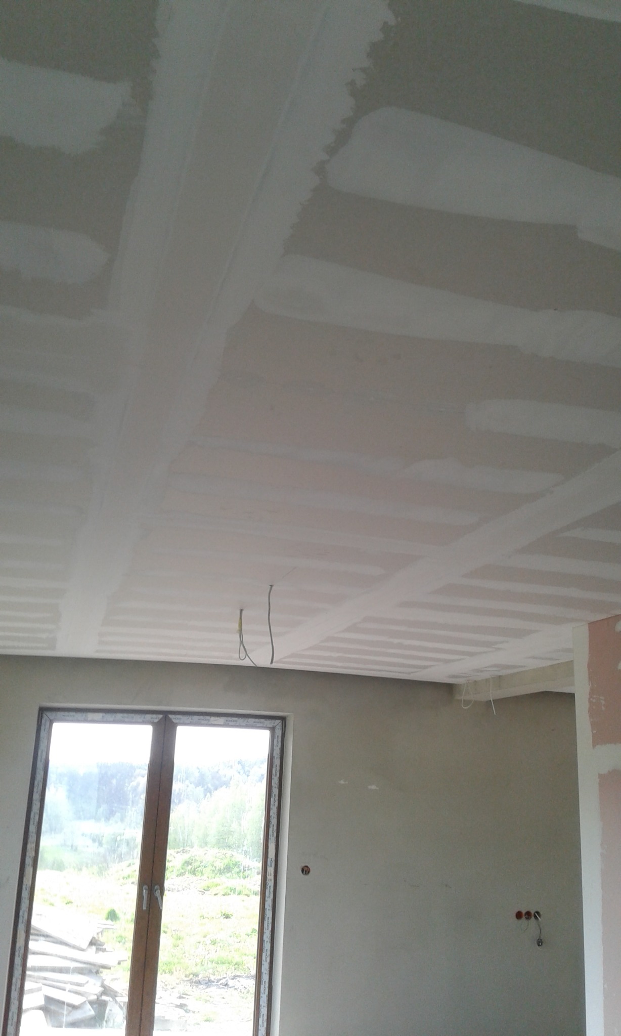 Sufit z płyt gipsowo-kartonowych w trakcie szpachlowania, widoczne łączenia płyt, fragment okna z widokiem na zewnątrz oraz wystające kable elektryczne.