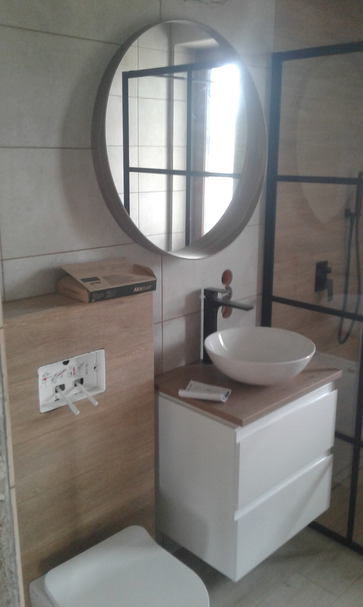 Nowoczesna łazienka w trakcie wykańczania: okrągłe lustro z drewnianą ramą, nablatowa umywalka z czarną baterią, podwieszana toaleta z zamontowanym stelażem, widoczna paczka Alca Plast.