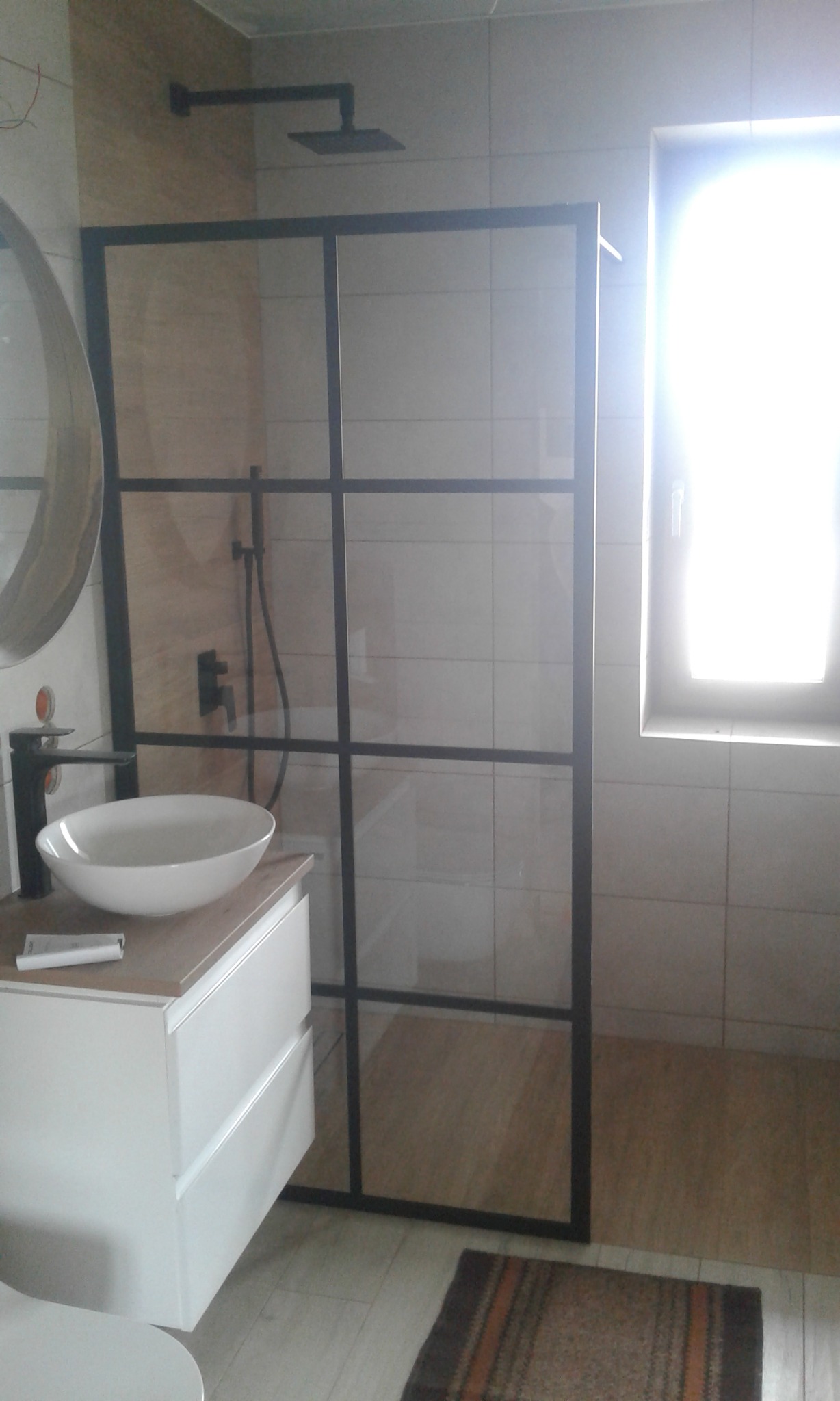 Nowoczesna łazienka z kabiną prysznicową w stylu loft z czarnymi profilami, białą umywalką nablatową i baterią w kolorze czarnym, oraz jasnymi płytkami na ścianach i podłodze. W tle widoczne okno...