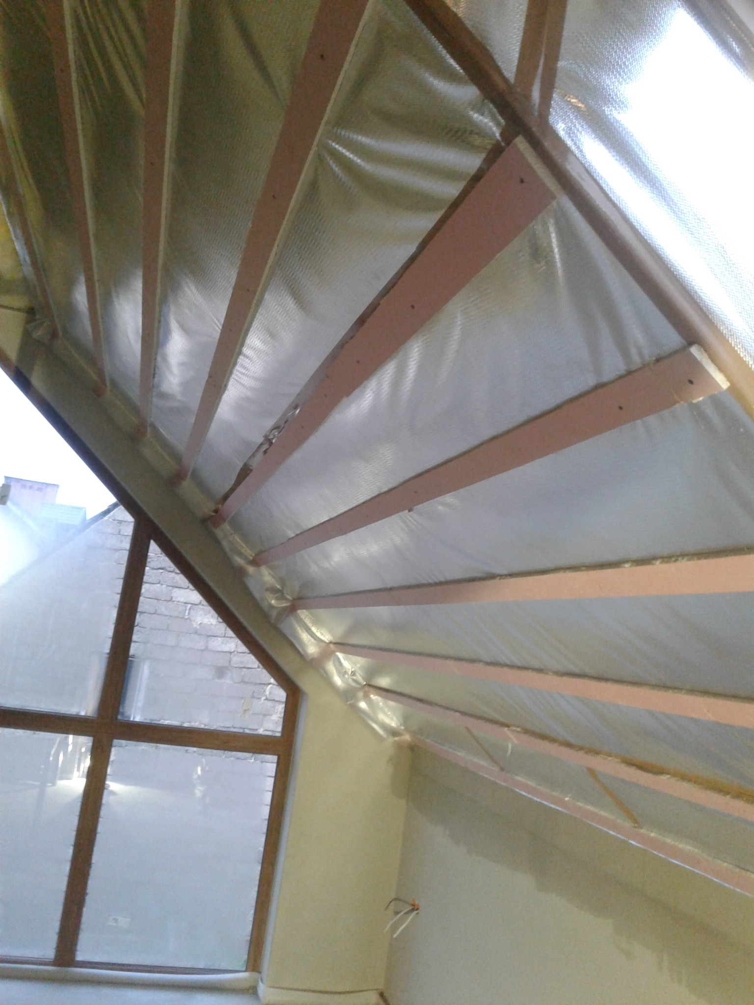 Skośny sufit poddasza z widoczną izolacją folią aluminiową i drewnianą konstrukcją nośną, okno w tle.
