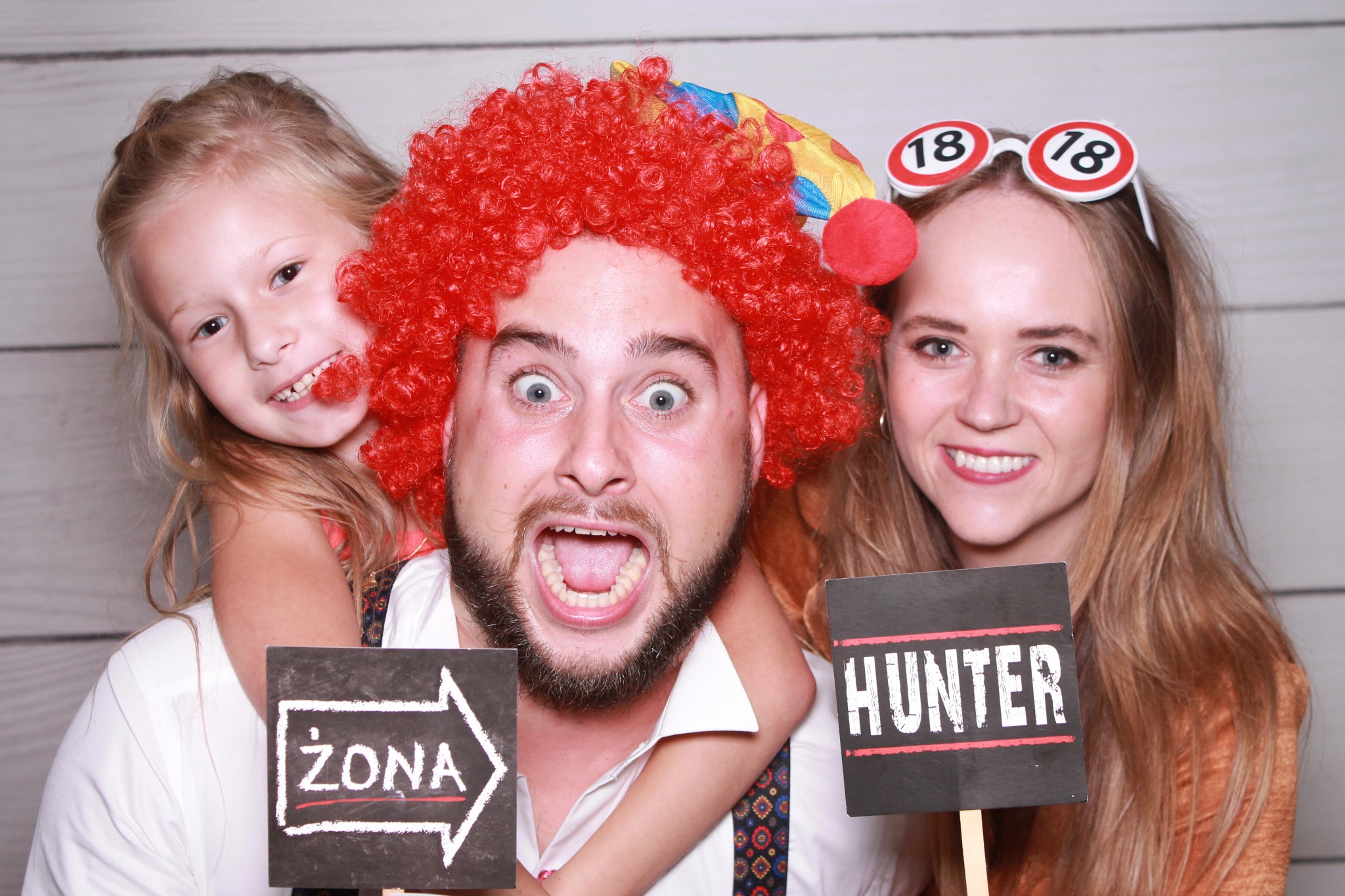 Zdjęcie z fotobudki: mężczyzna w czerwonej peruce z szeroko otwartymi ustami, trzymany przez dziewczynkę z napisem 'Żona', obok kobieta z okularami '18' i napisem 'Hunter'.
