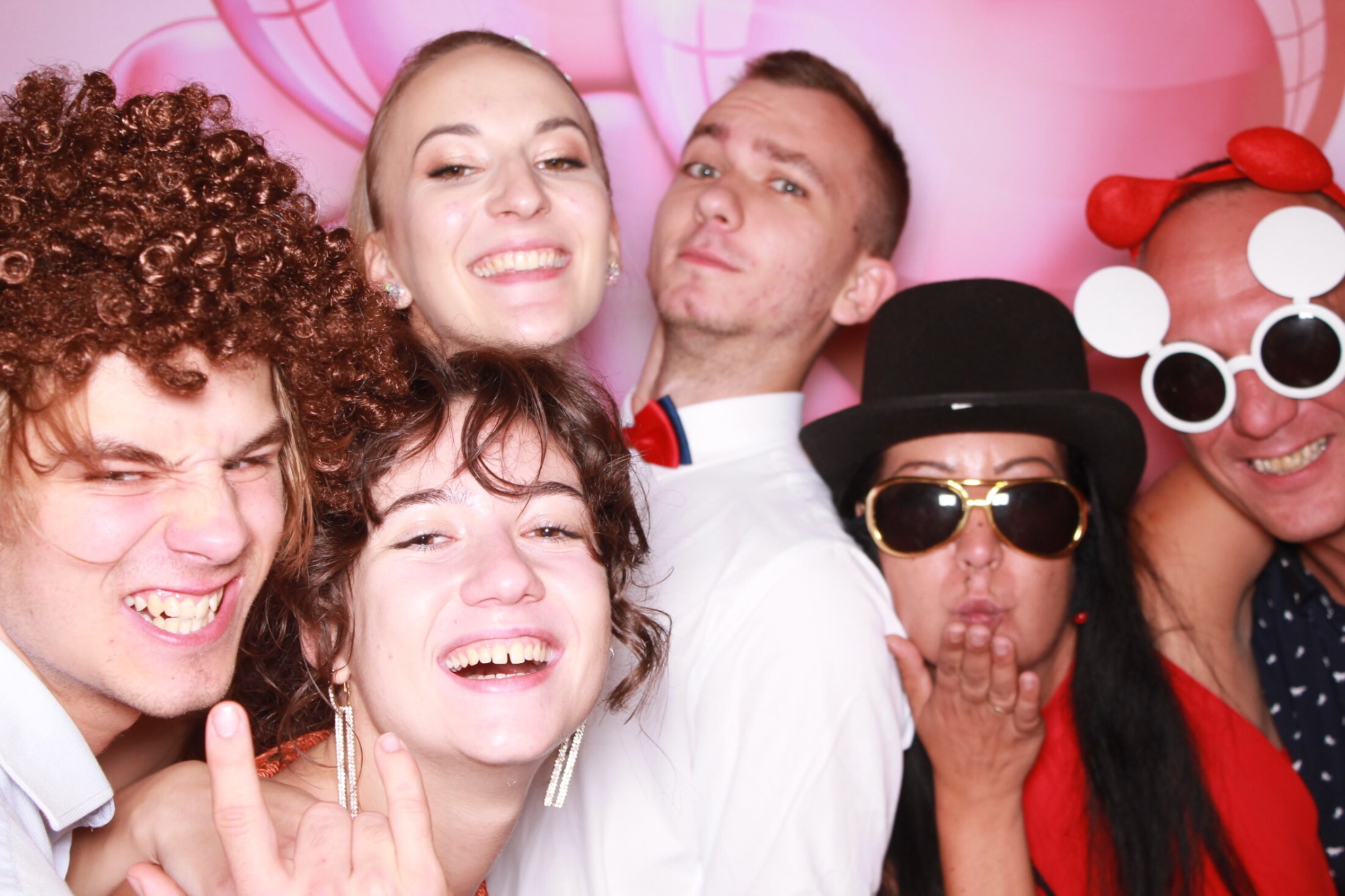 Grupa roześmianych osób pozuje do zdjęcia w fotobudce, ubrani w peruki, śmieszne okulary i inne rekwizyty na tle różowego tła z balonami.