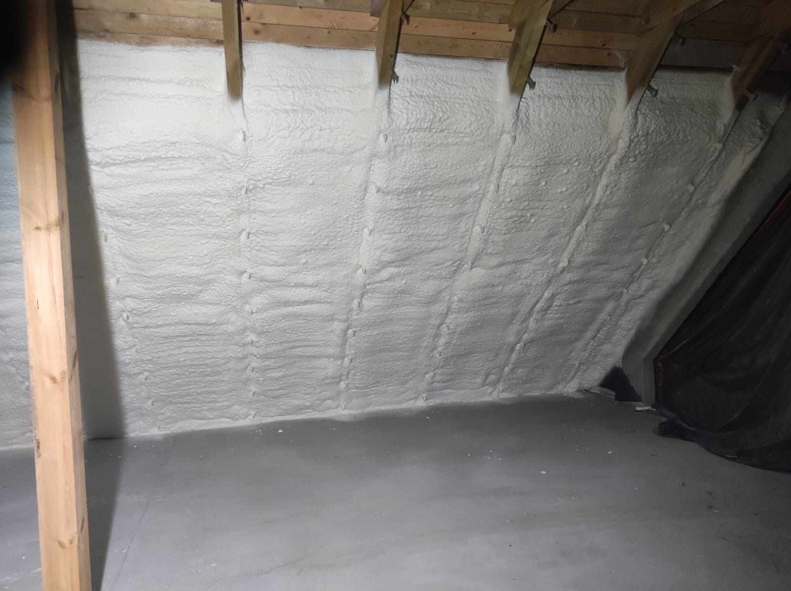 Poddasze po ociepleniu pianką PUR, widoczne drewniane elementy konstrukcyjne i szara podłoga. Izolacja równomiernie pokrywa skośne ściany.