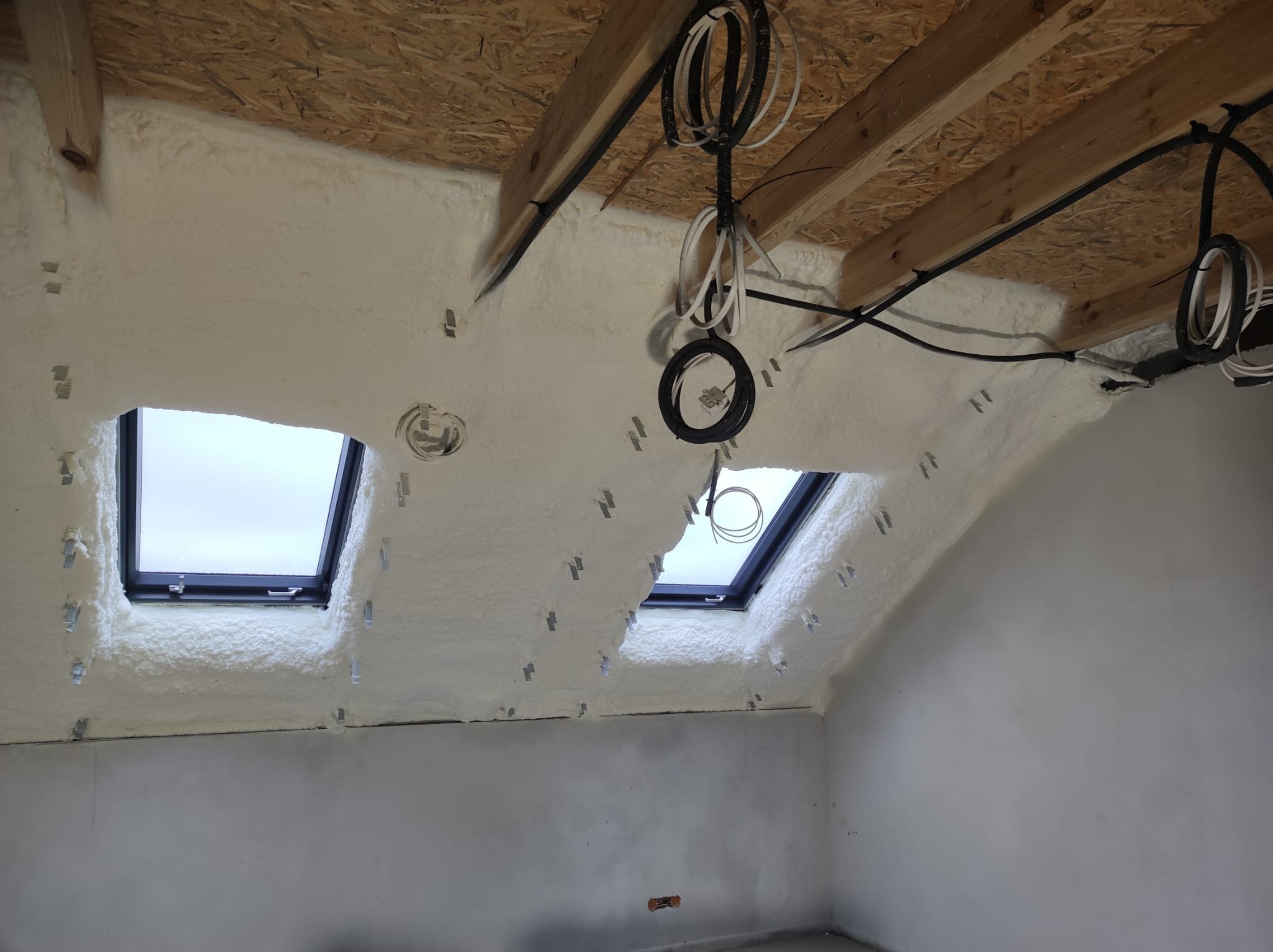 Poddasze z oknami dachowymi po aplikacji izolacji pianką PUR, widoczne kable elektryczne i elementy konstrukcji drewnianej. Biała, porowata struktura pianki.