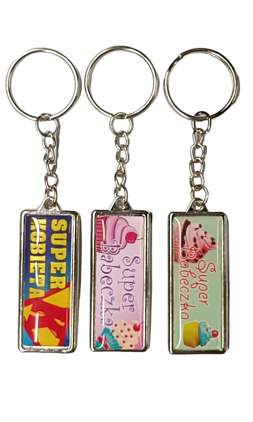 Trzy metalowe breloczki z grafikami: superbohaterki, różowego babeczki i zielonej babeczki, zawieszone na srebrnych kółkach i łańcuszkach, na białym tle.