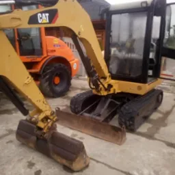 Żółta mini koparka CAT z łyżką i lemieszem na betonowym podłożu, w tle pomarańczowa maszyna budowlana i dom. Widoczne ślady użytkowania.