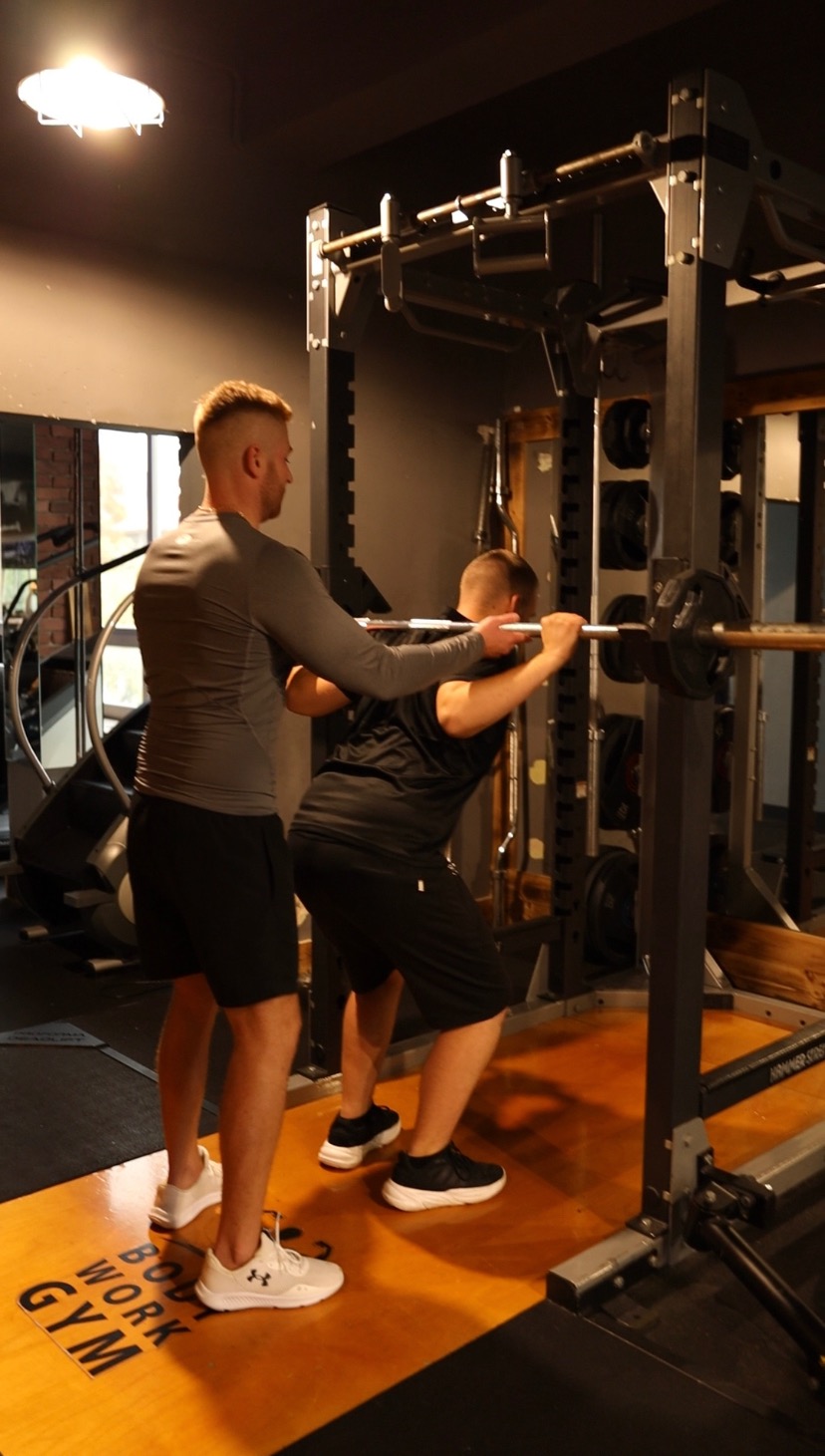 Mężczyzna w szarej koszulce asekuruje mężczyznę wykonującego przysiad ze sztangą w siłowni. Na podłodze napis BODY WORK GYM.