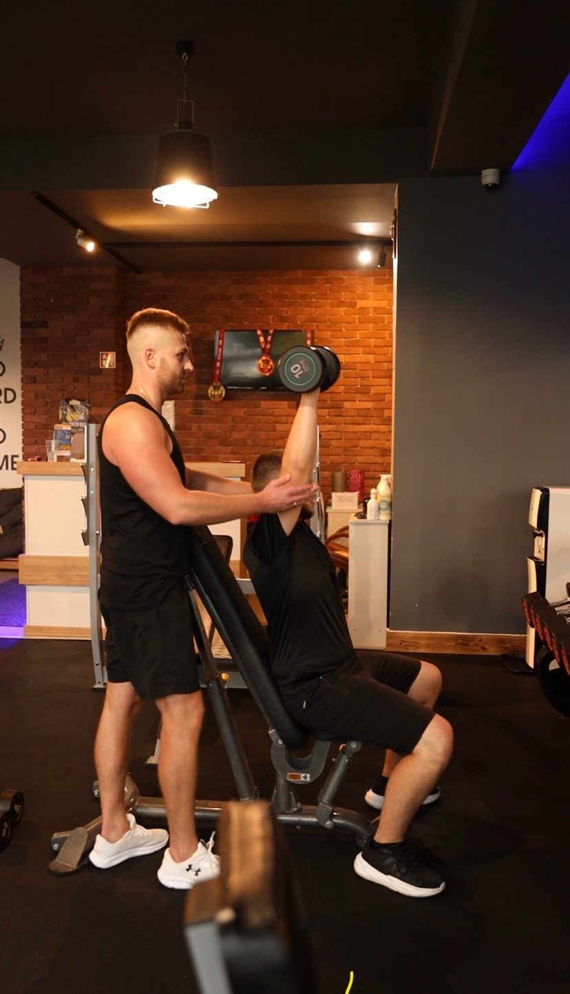 Mężczyzna asekuruje ćwiczącego na ławce skośnej z hantlą nad głową w siłowni, widoczna ceglana ściana i wiszące medale.