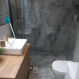 Nowoczesna łazienka z szarymi marmurowymi płytkami, złotą armaturą i drewnianą szafką pod umywalką. Widoczny mop oparty o ścianę w kabinie prysznicowej sugerujący świeżo zakończone prace porządkowe.