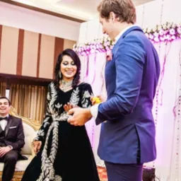 Uśmiechnięta panna młoda w czarnej sukni z srebrnymi zdobieniami, mężczyzna w niebieskim garniturze wręcza jej mały przedmiot, w tle siedzący mężczyzna w smokingu i dekoracja kwiatowa.