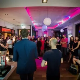 Iluzjonista w niebieskiej marynarce prezentuje sztuczkę przed zgromadzonym tłumem w oświetlonym pomieszczeniu z dekoracyjnymi kulami pod sufitem, widoczny otwarty kufer z rekwizytami.