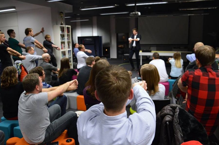 Publiczność siedzi na kolorowych pufach, obserwując występującego na scenie mężczyznę w garniturze, który wykonuje gest ręką, a w tle widać biały stolik i duży ekran projekcyjny.