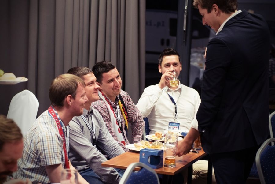 Grupa mężczyzn w strojach biznesowych siedzi przy stole z przekąskami i napojami, jeden z nich pije piwo, a drugi w garniturze stoi obok, możliwe, że w trakcie przerwy konferencyjnej.