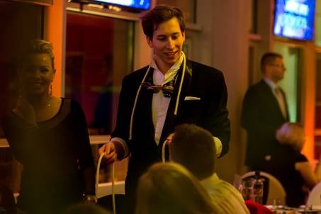 Młody mężczyzna w smokingu z muszką na szyi, wykonujący trik z linami na tle sali bankietowej z gośćmi.