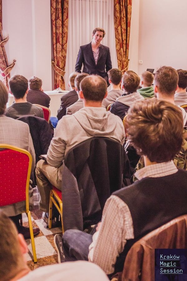 Prezentacja na temat iluzji: mężczyzna w garniturze na scenie przed publicznością siedzącą na czerwonych krzesłach, widok z tyłu sali.