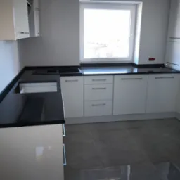 Nowoczesna kuchnia z białymi szafkami i czarnym blatem, widoczny fragment instalacji elektrycznej na ścianie.