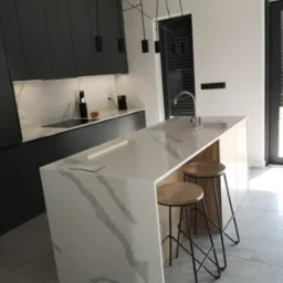 Nowoczesna kuchnia z wyspą i blatem z konglomeratu kwarcowego z widocznymi żyłami, oświetlona wiszącymi lampami i podświetleniem LED.