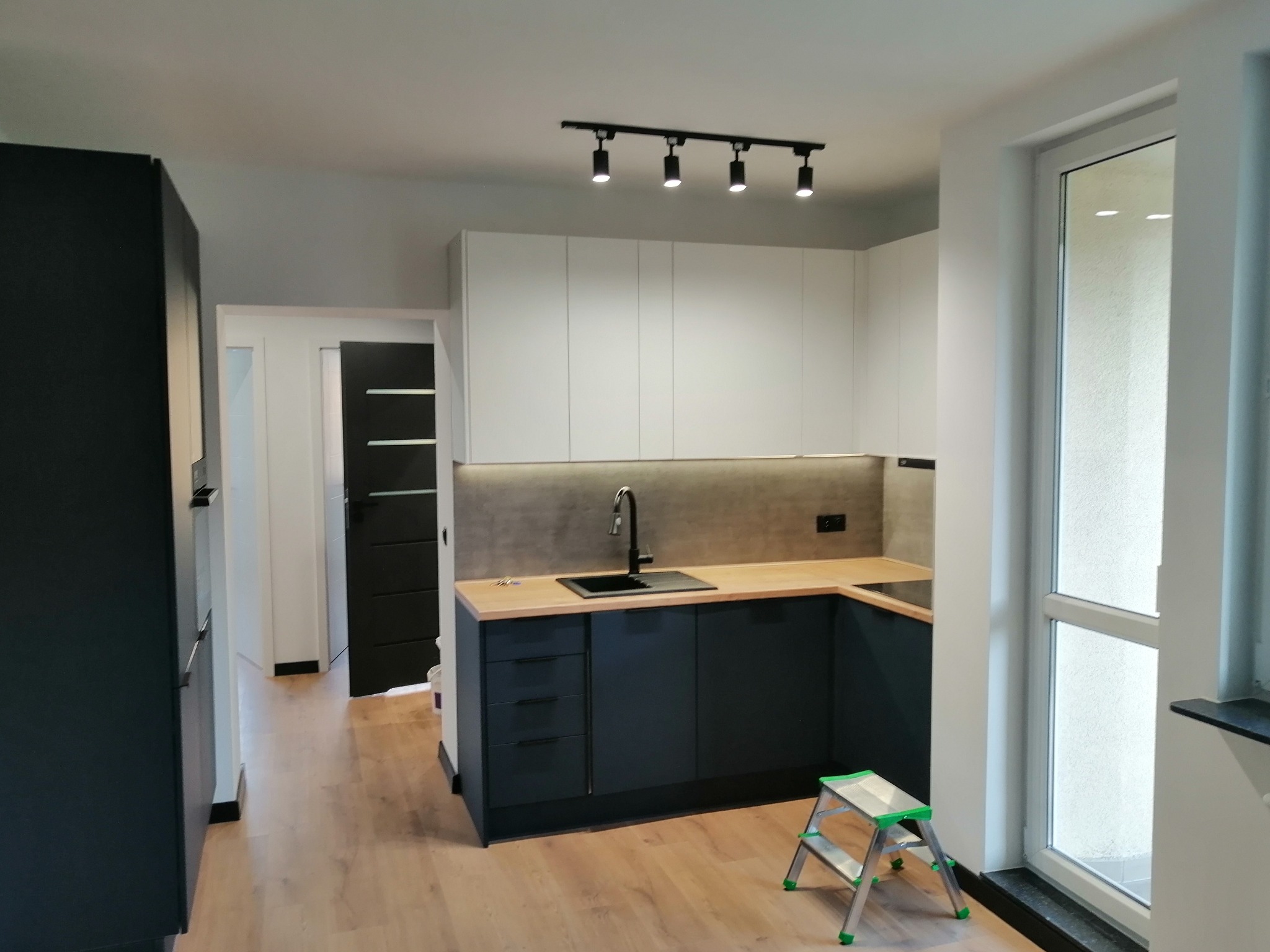 Nowoczesna kuchnia z czarnymi szafkami dolnymi i jasnymi górnymi, drewnianym blatem i czarnym zlewem, oświetlona reflektorami, z małą drabinką w rogu.