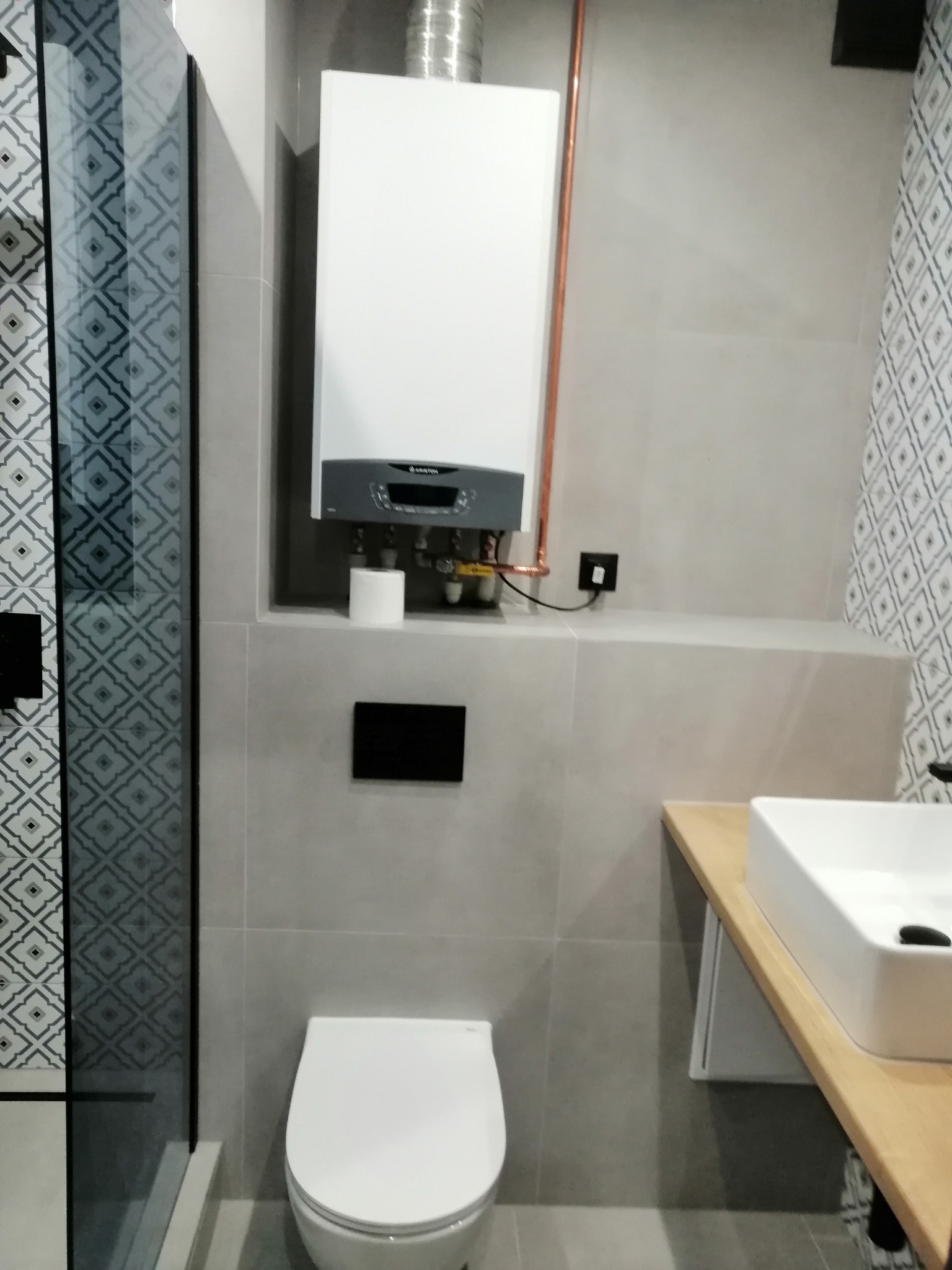 Nowoczesna łazienka z szarymi płytkami, kabiną prysznicową z wzorzystą ścianą, wiszącą toaletą, umywalką na drewnianym blacie i zamontowanym piecem gazowym Ariston.