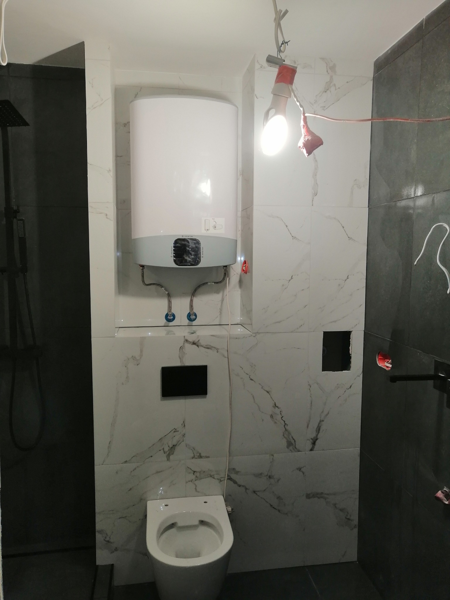 Biały podgrzewacz wody Ariston zamontowany na ścianie z marmurowymi płytkami, widoczna toaleta, czarna kabina prysznicowa i prowizoryczne oświetlenie budowlane w trakcie prac.