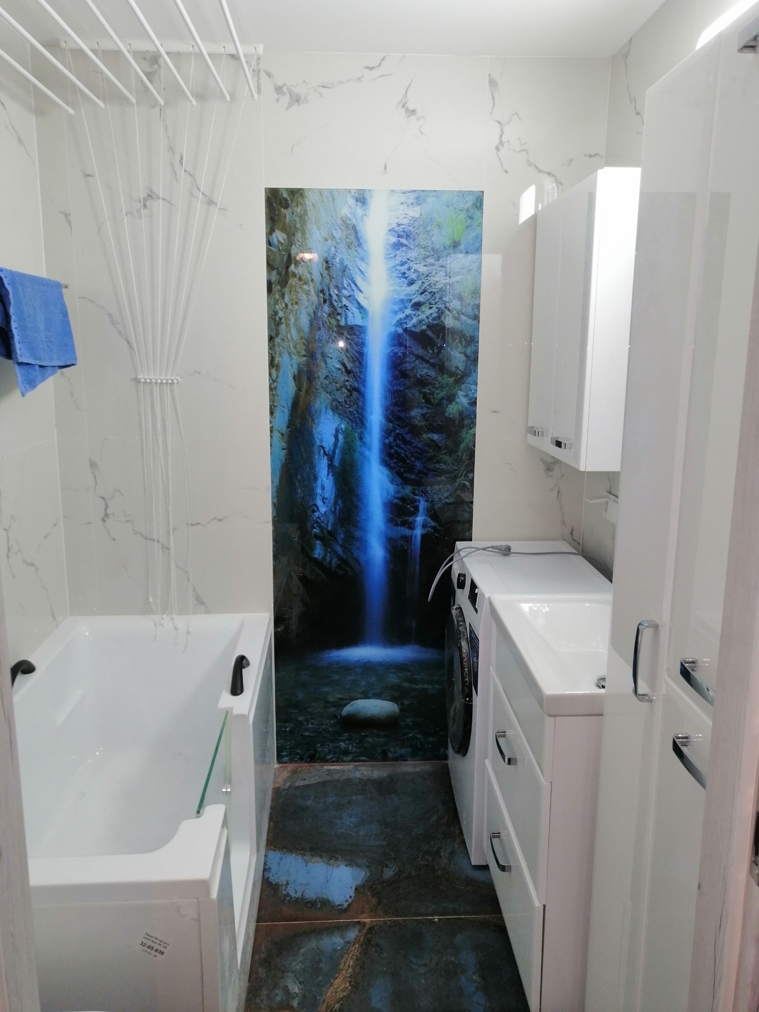 Biała łazienka z wanną, pralką i szafkami, wyróżniająca się panelem ściennym z motywem wodospadu, oraz suszarką sufitową na pranie.