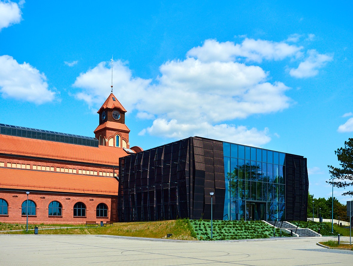 Widok zewnętrzny budynku z ceglaną wieżą zegarową i nowoczesną szklaną fasadą z drewnianymi elementami, jasne niebo z chmurami w tle.
