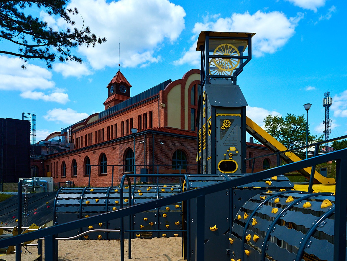 Plac zabaw z motywem kopalni na tle odrestaurowanego budynku z czerwonej cegły z wieżą zegarową, niebieskie niebo z chmurami w tle.