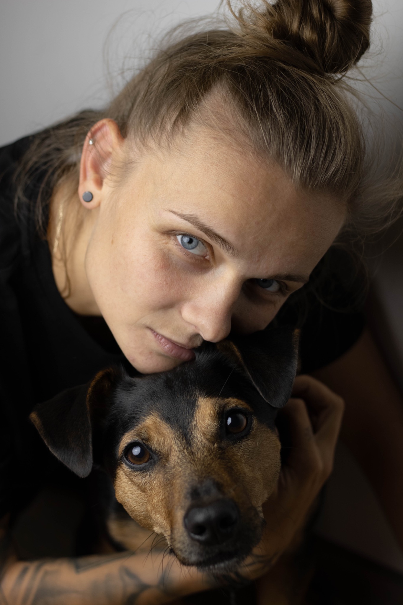 Portret kobiety o niebieskich oczach, z włosami upiętymi w kok, obejmującej czarno-brązowego psa rasy pinczer miniaturowy, widoczne tatuaże na rękach.