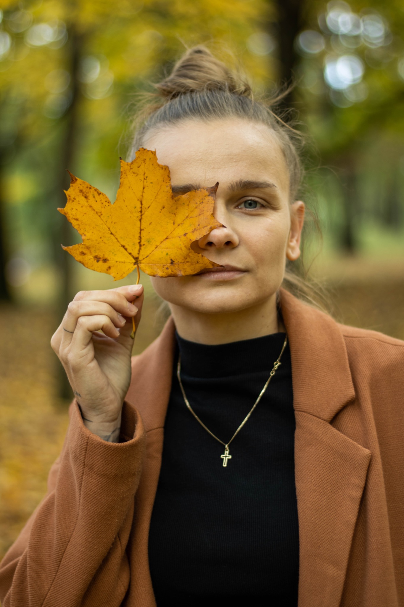 Portret kobiety z jesiennym liściem klonu zakrywającym część twarzy, w tle park z opadłymi liśćmi.