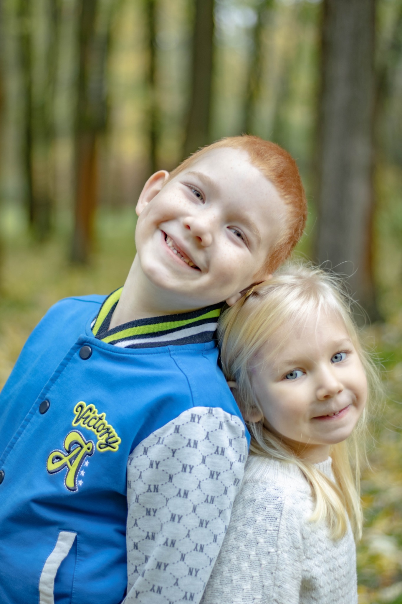 Uśmiechnięty rudowłosy chłopiec w niebieskiej kurtce z napisem Victory opiera głowę o blondwłosą dziewczynkę na tle rozmytego lasu.