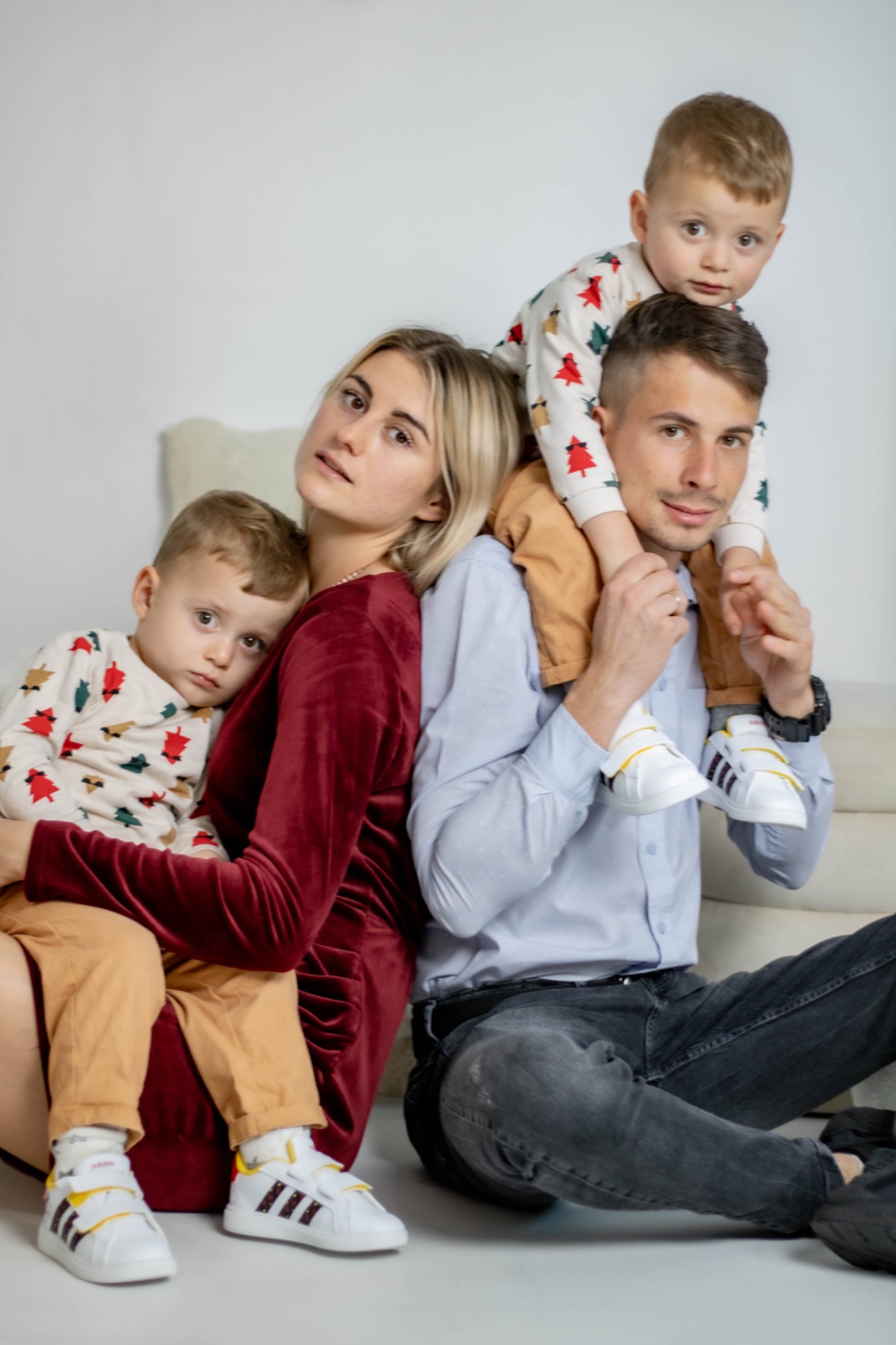 Portret rodziny: mama w bordowej sukience, tata w błękitnej koszuli, syn siedzący na kolanach mamy, syn na barana u taty, wszyscy w świątecznych strojach i białych adidasach, na jasnym tle.