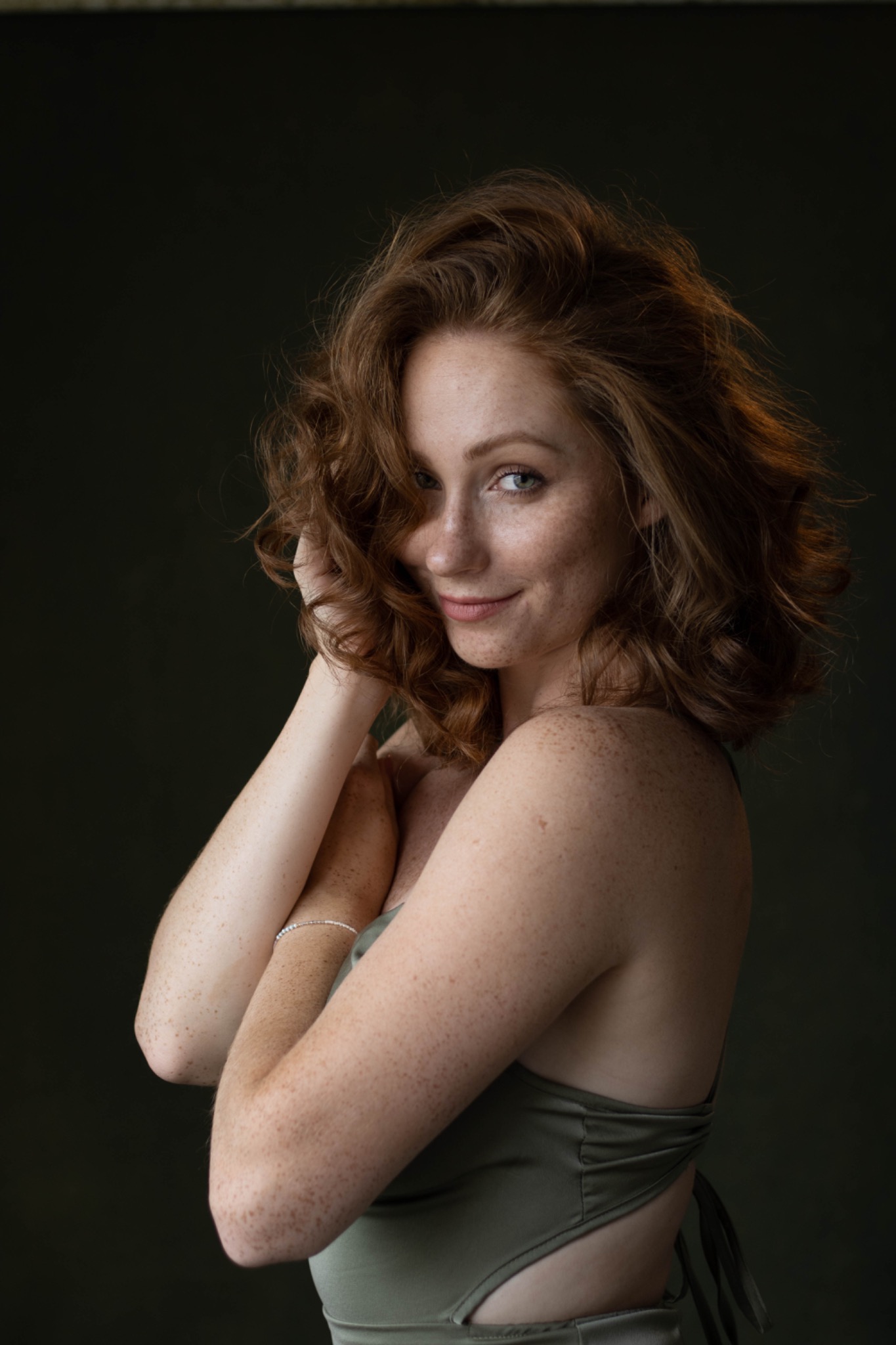 Portret uśmiechniętej kobiety z rudymi, kręconymi włosami i piegami, pozującej na ciemnym tle w oliwkowym topie z odkrytymi plecami i delikatną bransoletką.