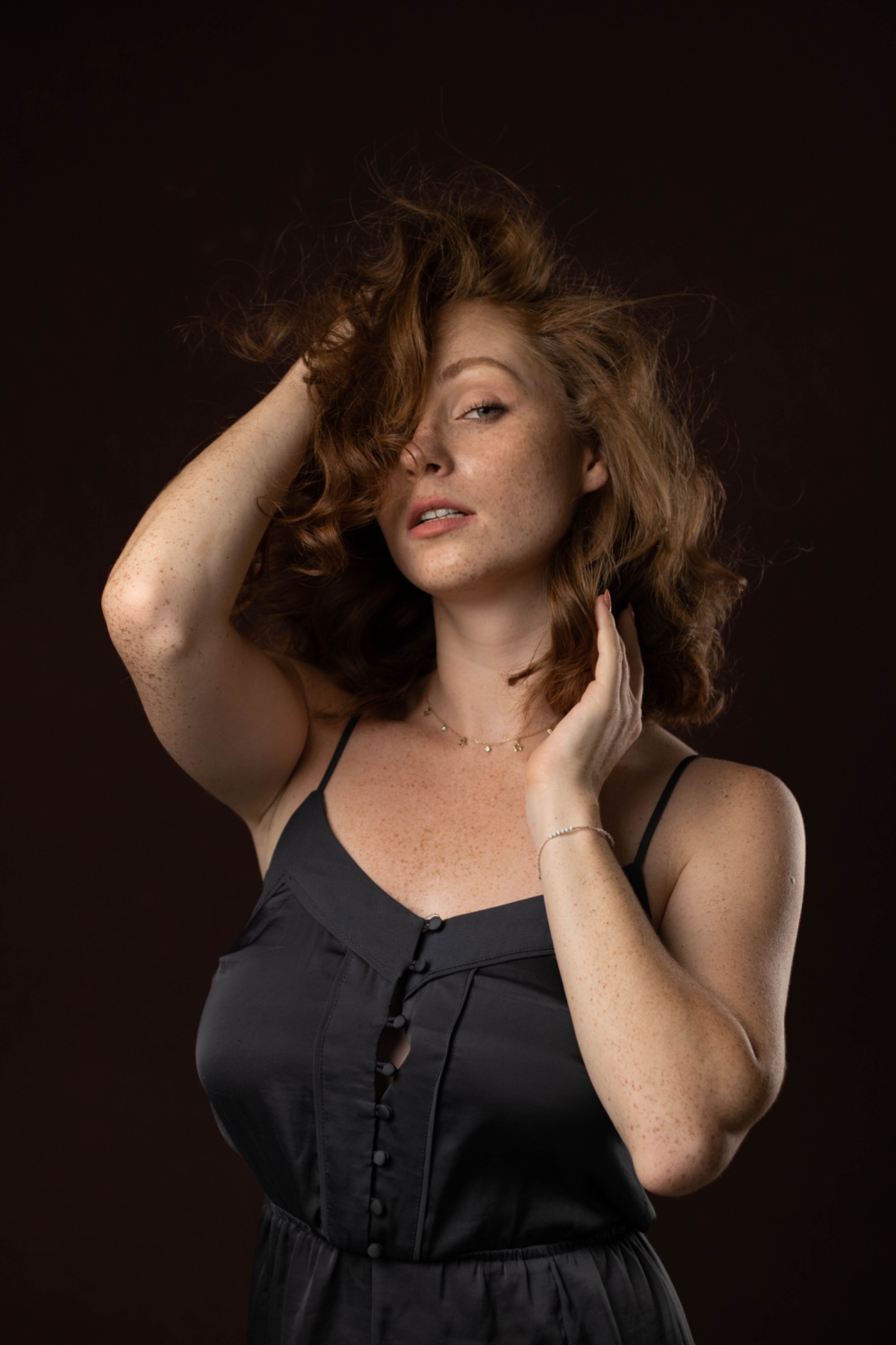Portret kobiety z rudymi włosami i piegami, ubrana w czarną satynową sukienkę na ramiączkach, pozująca na ciemnobrązowym tle.