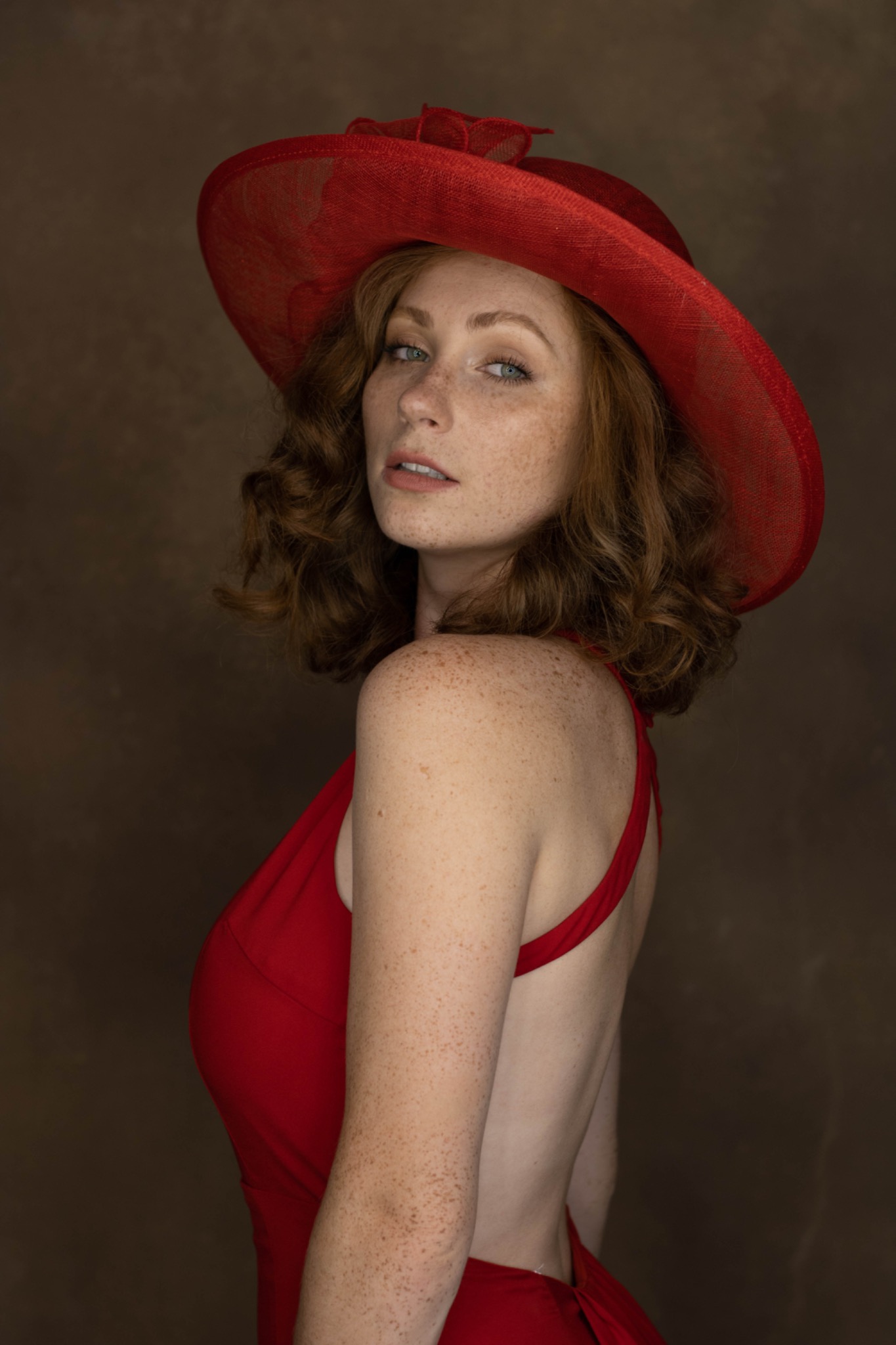 Portret kobiety z rudymi kręconymi włosami i piegami, ubrana w czerwoną sukienkę i kapelusz, spoglądająca przez ramię na brązowym tle.