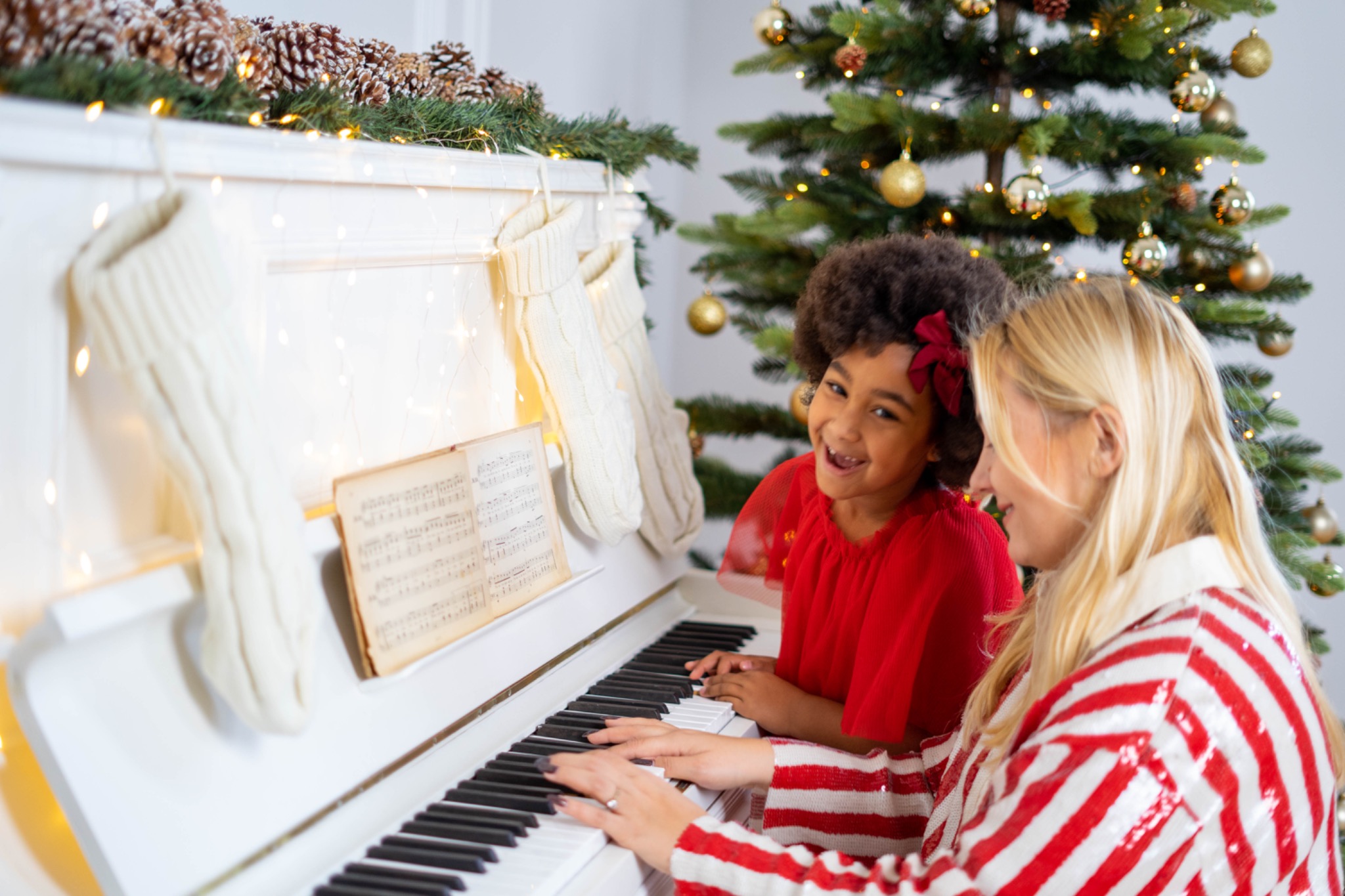 Kobieta gra na białym pianinie, obok stoi uśmiechnięta dziewczynka; w tle choinka i świąteczne dekoracje.