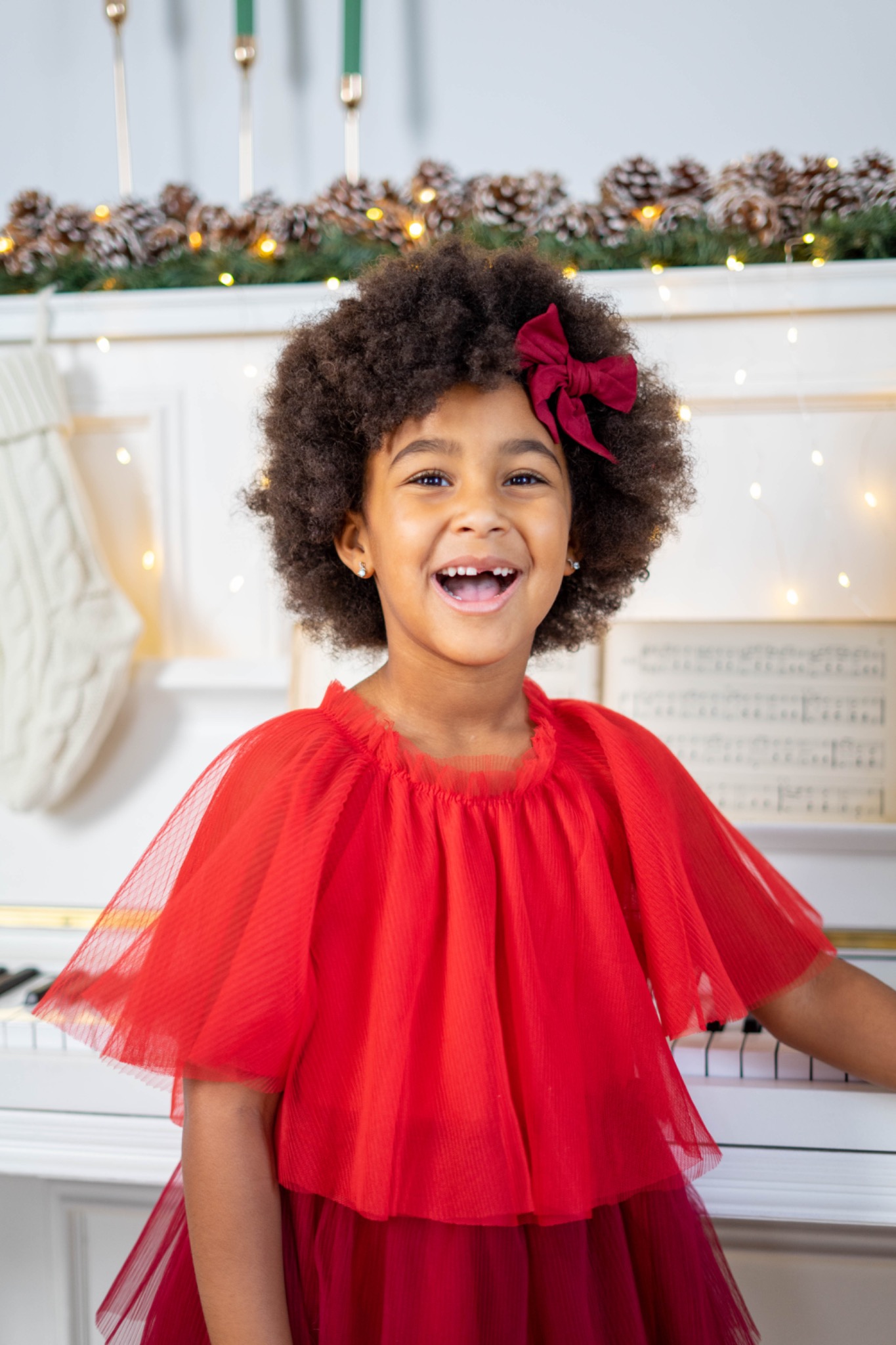 Uśmiechnięta dziewczynka z afro i czerwoną kokardą we włosach, ubrana w czerwoną sukienkę z tiulu, stoi przy białym pianinie, w tle świąteczna dekoracja z szyszek i światełek.