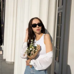 Kobieta w okularach przeciwsłonecznych, opierająca się o kolumnę, trzymająca bukiet w papierowym opakowaniu, w tle widoczne okno.