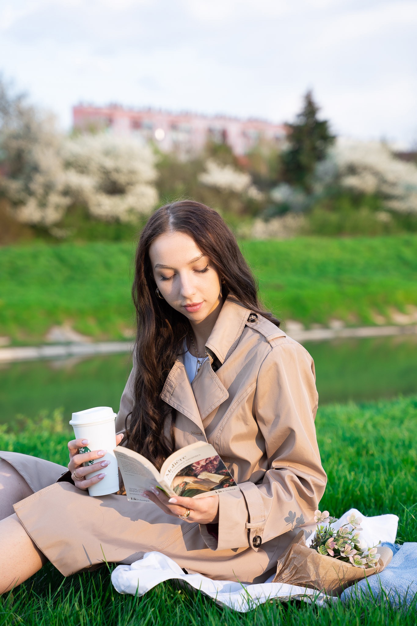 Kobieta w beżowym płaszczu siedzi na trawie nad rzeką, czyta książkę i trzyma kubek z kawą, obok leżą kwiaty.