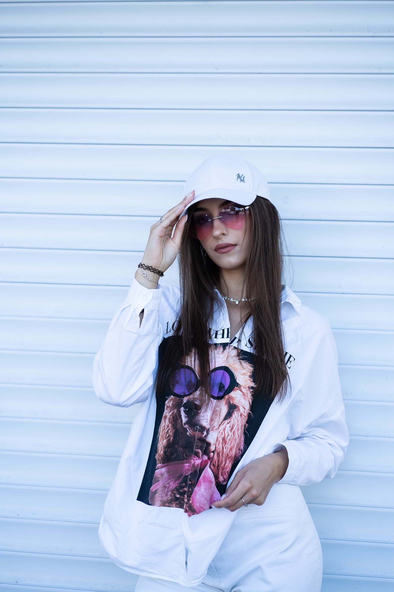 Kobieta w białym stroju pozuje na tle białej rolety, ma na sobie koszulkę z nadrukiem lamy w okularach i różowe okulary w kształcie serc.