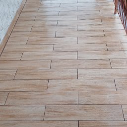 ART-REMONT Robert Skrzypczak - Balkon wyłożony podłużnymi płytkami imitującymi drewno, z metalową balustradą i widokiem na zieleń.