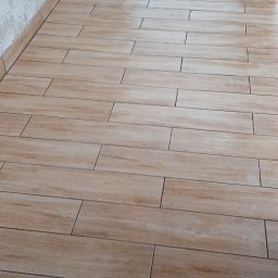 ART-REMONT Robert Skrzypczak - Beżowe płytki podłogowe imitujące drewno na balkonie, z widoczną teksturą drewna i fugami, ściana z białej cegły dekoracyjnej w tle.